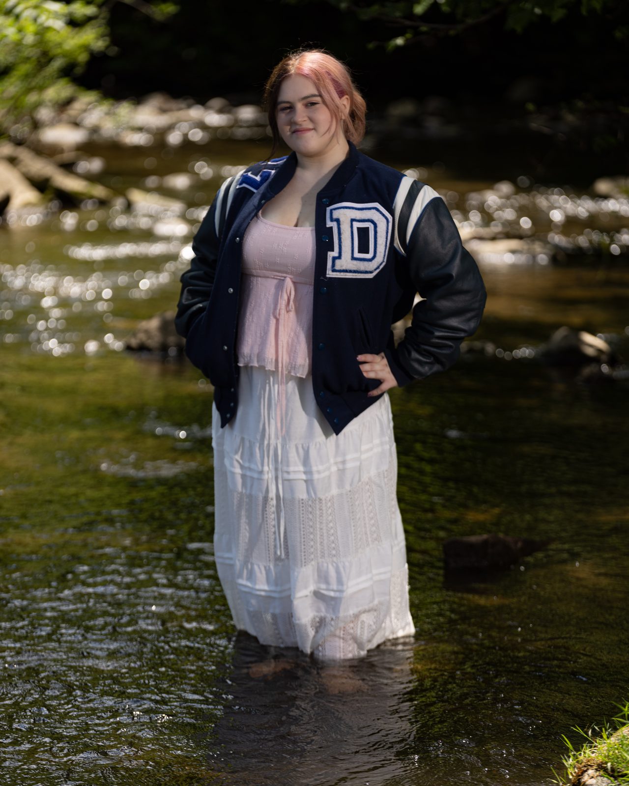 High school senior in letterman jacket and floral dress by a stream — Ann Arbor senior portraits